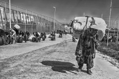 A woman walks towards the border with a heavy pack on her back, while a group of men push tires. Melilla. Spain